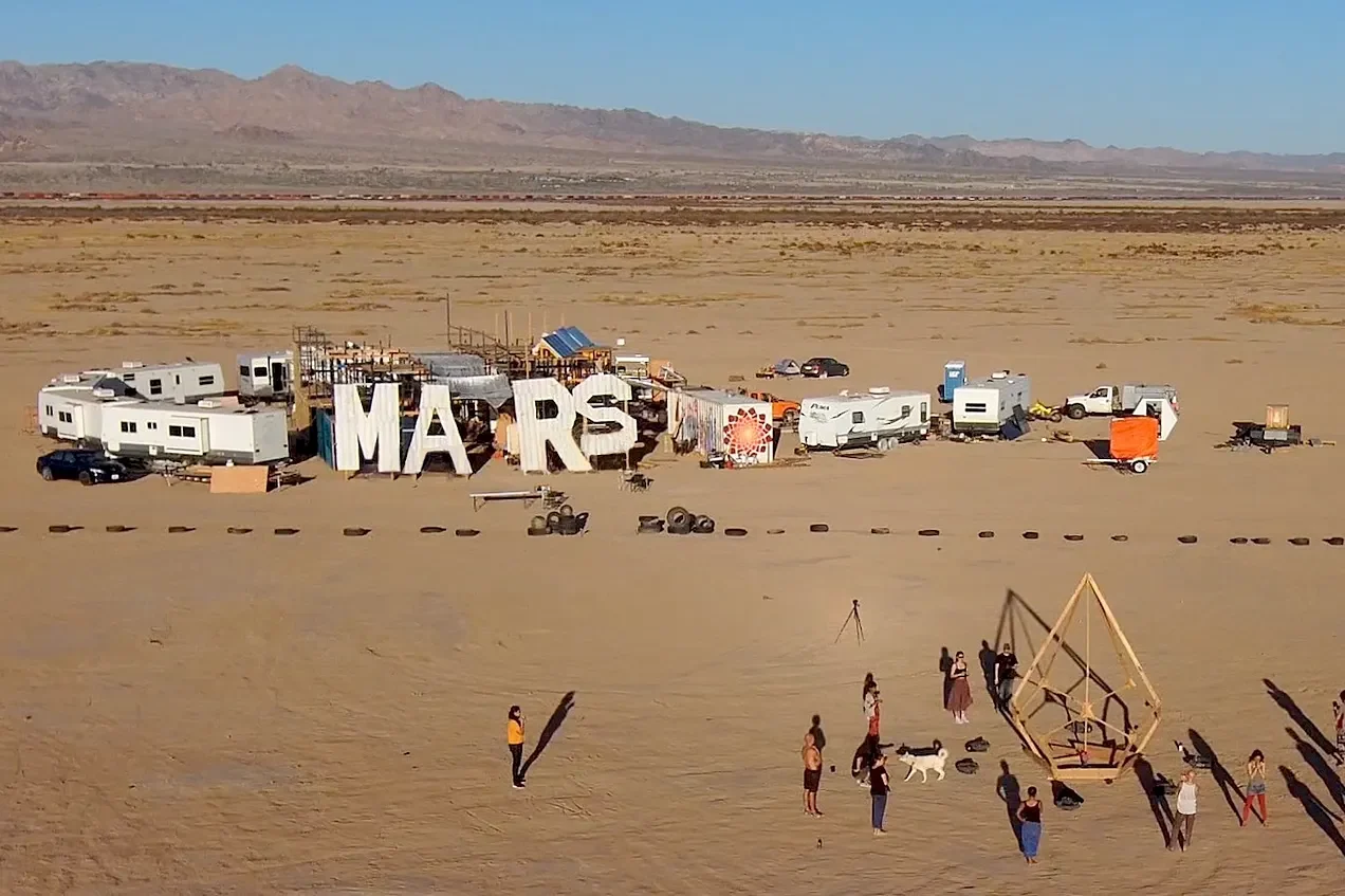 The camp from the air: the MARS letters, the trailers and the pallet-rack building on the dry lake bed, with people gathered around a wooden frame in the foreground.