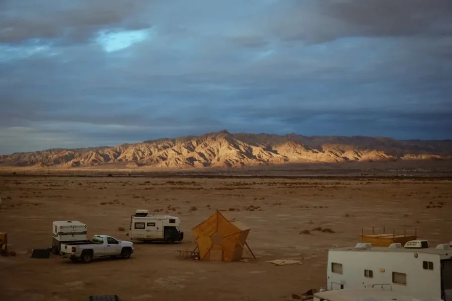 The Mars College camp on the dry lake bed at dusk: the solar array, the pallet-rack building and the vans parked around it.