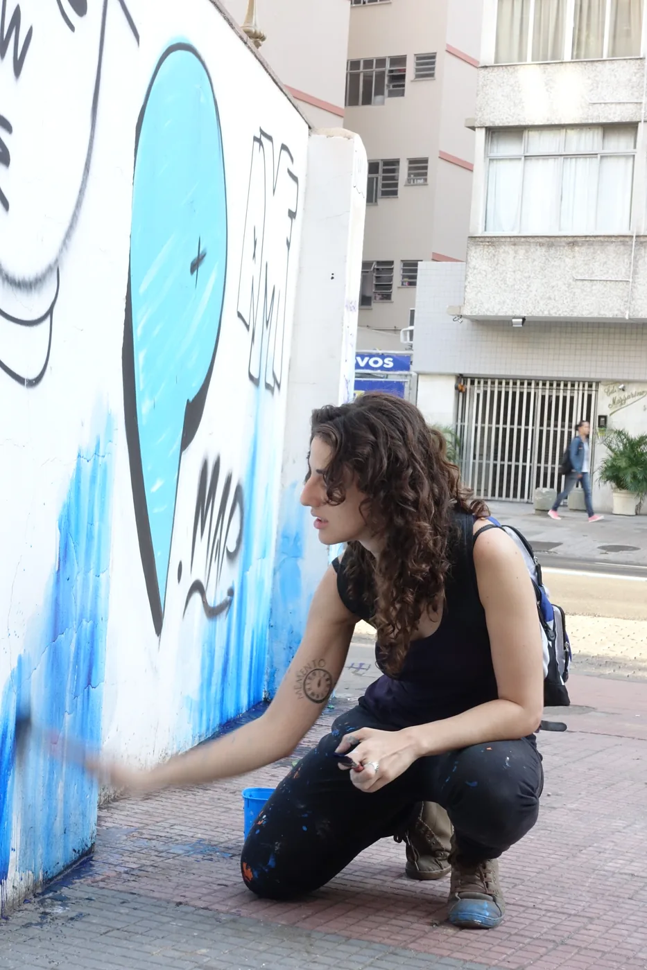 Vanessa Rosa crouching on a pavement, painting blue onto a street wall with a brush.