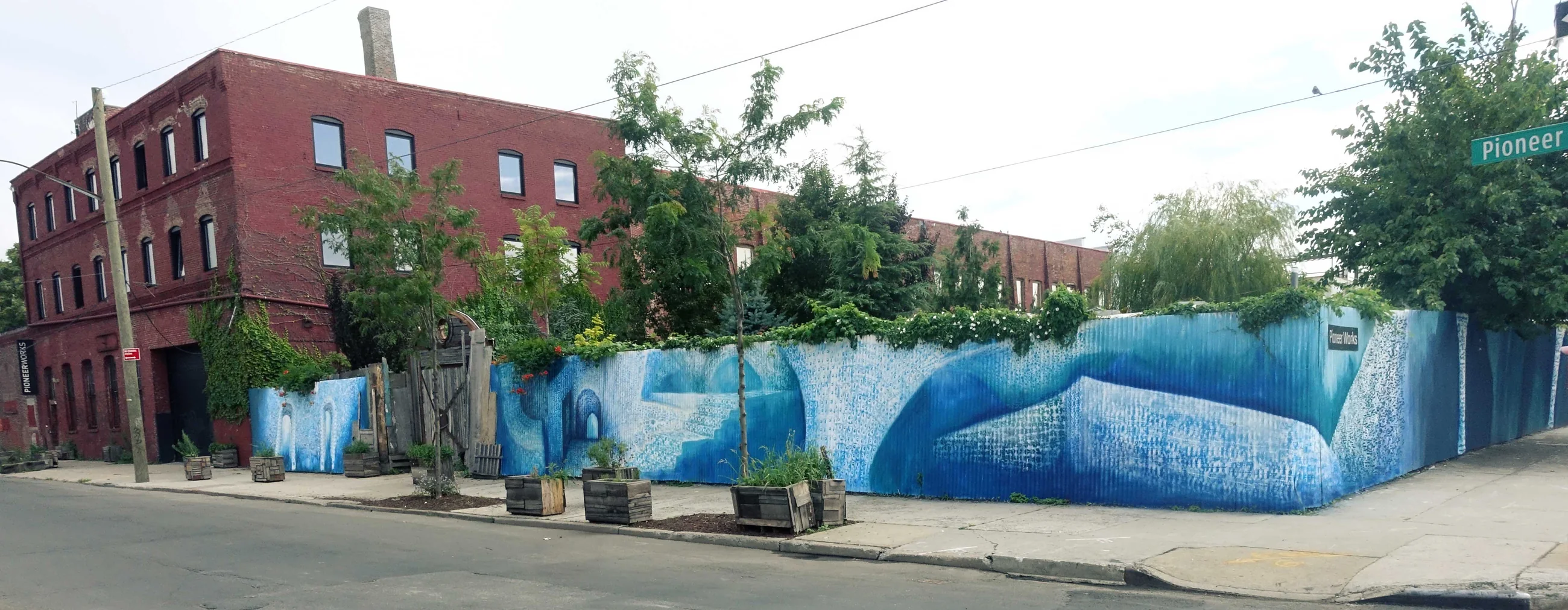 The painted blue fence wrapping the corner of Pioneer Works in Red Hook, with the red brick building behind it.