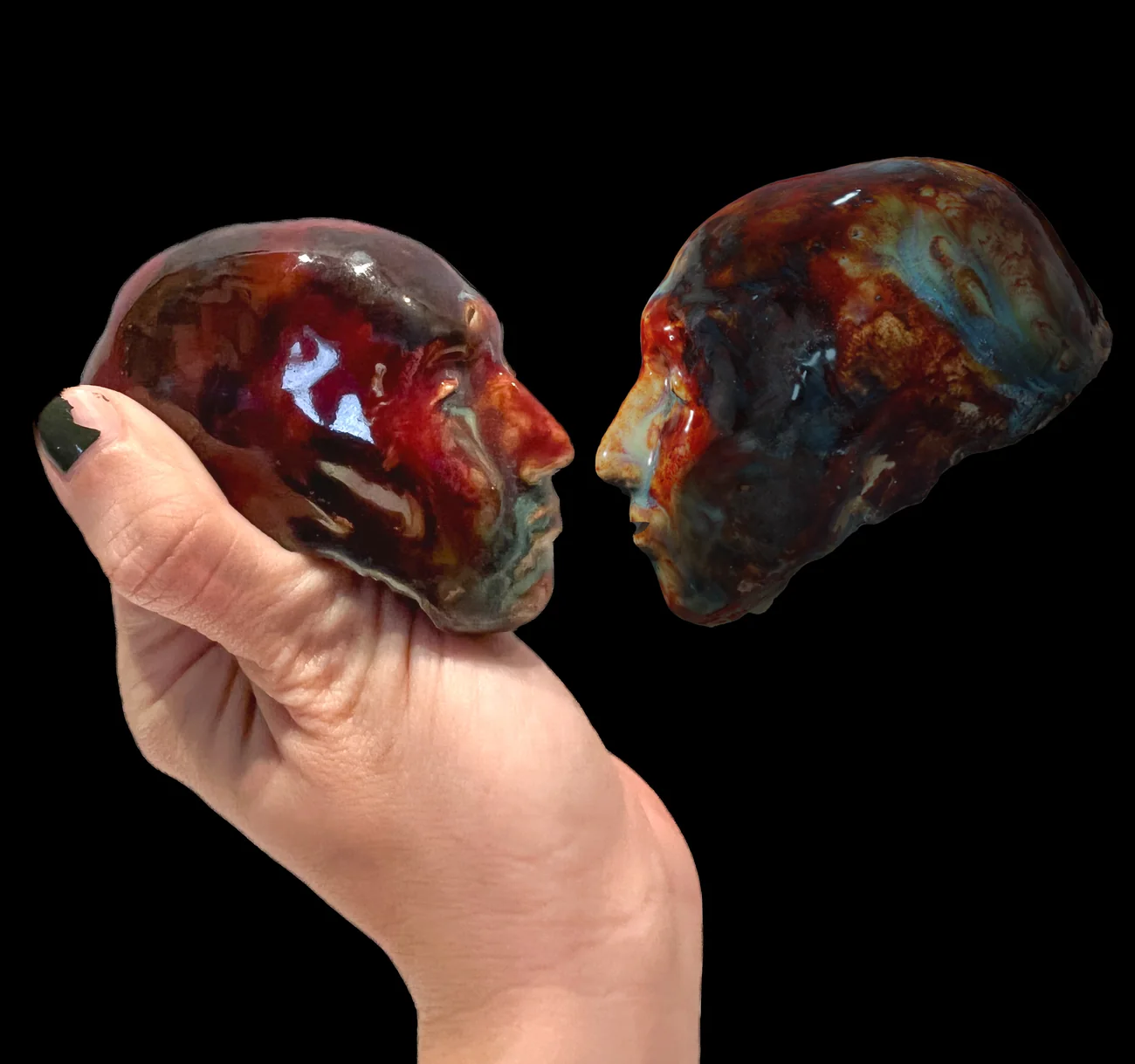 Two glazed ceramic Little Martian heads facing each other, one held in a hand, against black.