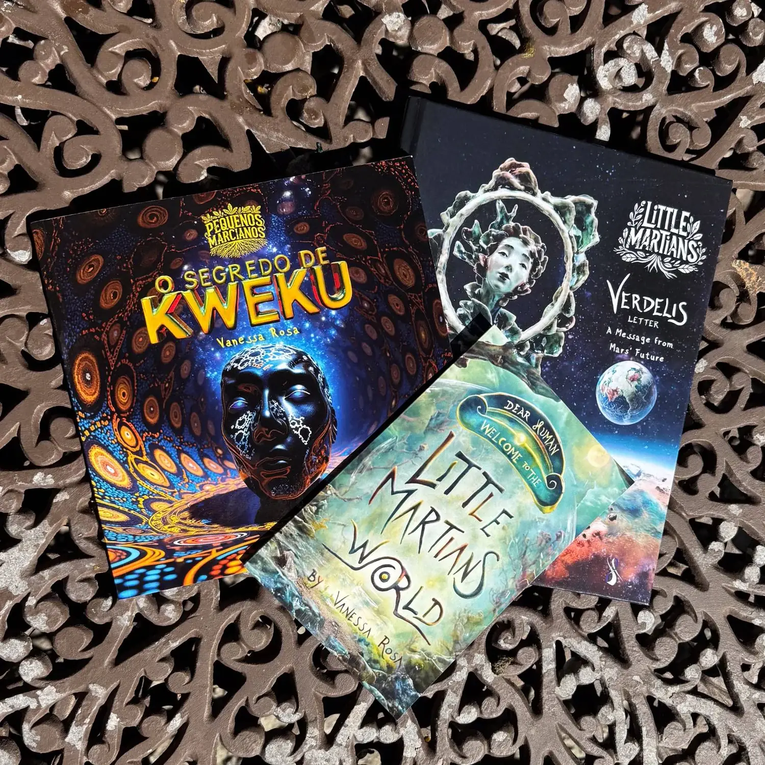 Three Little Martians books in print on a wrought-iron table: O Segredo de Kweku, Verdelis Letter, and Dear Human, Welcome to the Little Martians World.